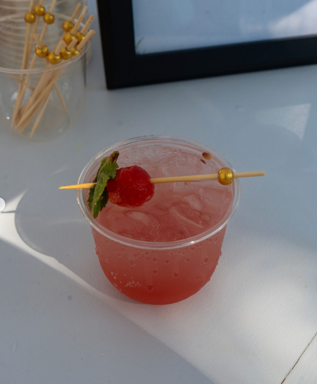 Iced pink cocktail in a clear plastic cup topped with a cherry and mint on a gold-beaded skewer, sitting on a white table.