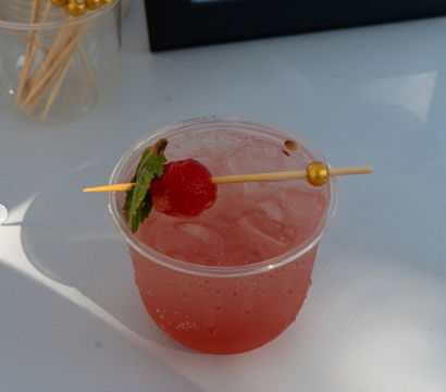 Iced pink cocktail in a clear plastic cup topped with a cherry and mint on a gold-beaded skewer, sitting on a white table.