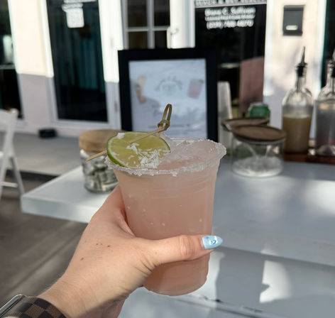 Sunlit hand holding a pink frozen cocktail in a salt-rimmed plastic cup with a lime wheel garnish at an outdoor patio bar.