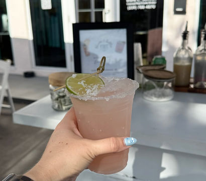 Sunlit hand holding a pink frozen cocktail in a salt-rimmed plastic cup with a lime wheel garnish at an outdoor patio bar.