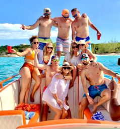 Nine friends on a boat in clear turquoise water near a tree-lined shore, wearing swimsuits, hats and sunglasses, smiling and holding drinks on a sunny tropical day.