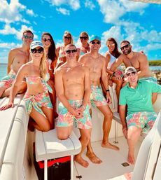 Cheerful group of friends on a motorboat in clear turquoise water, wearing matching colorful tropical-print swimwear and sunglasses under a bright blue sky on a sunny vacation day.