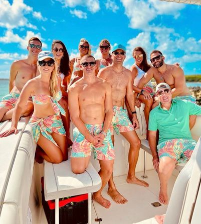 Cheerful group of friends on a motorboat in clear turquoise water, wearing matching colorful tropical-print swimwear and sunglasses under a bright blue sky on a sunny vacation day.