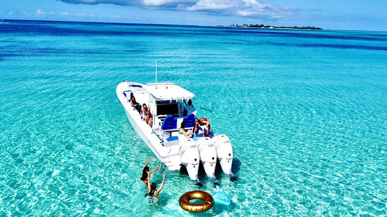 White powerboat with three outboard engines anchored in crystal-clear turquoise tropical water near a small island horizon; people lounge on the boat while two swimmers reach for a gold inflatable ring under a sunlit blue sky.