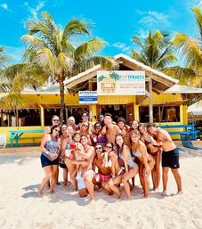 Large group of people posing on a white-sand tropical beach in front of a colorful yellow beach bar with palm trees and bright blue sky, holding drinks — fun summer vacation scene.