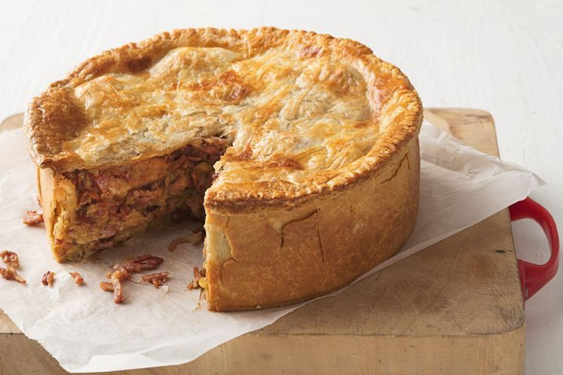 Mouthwatering deep-dish meat pie with a flaky golden crust and shredded meat filling, a wedge removed to reveal layered interior on a parchment-lined wooden board