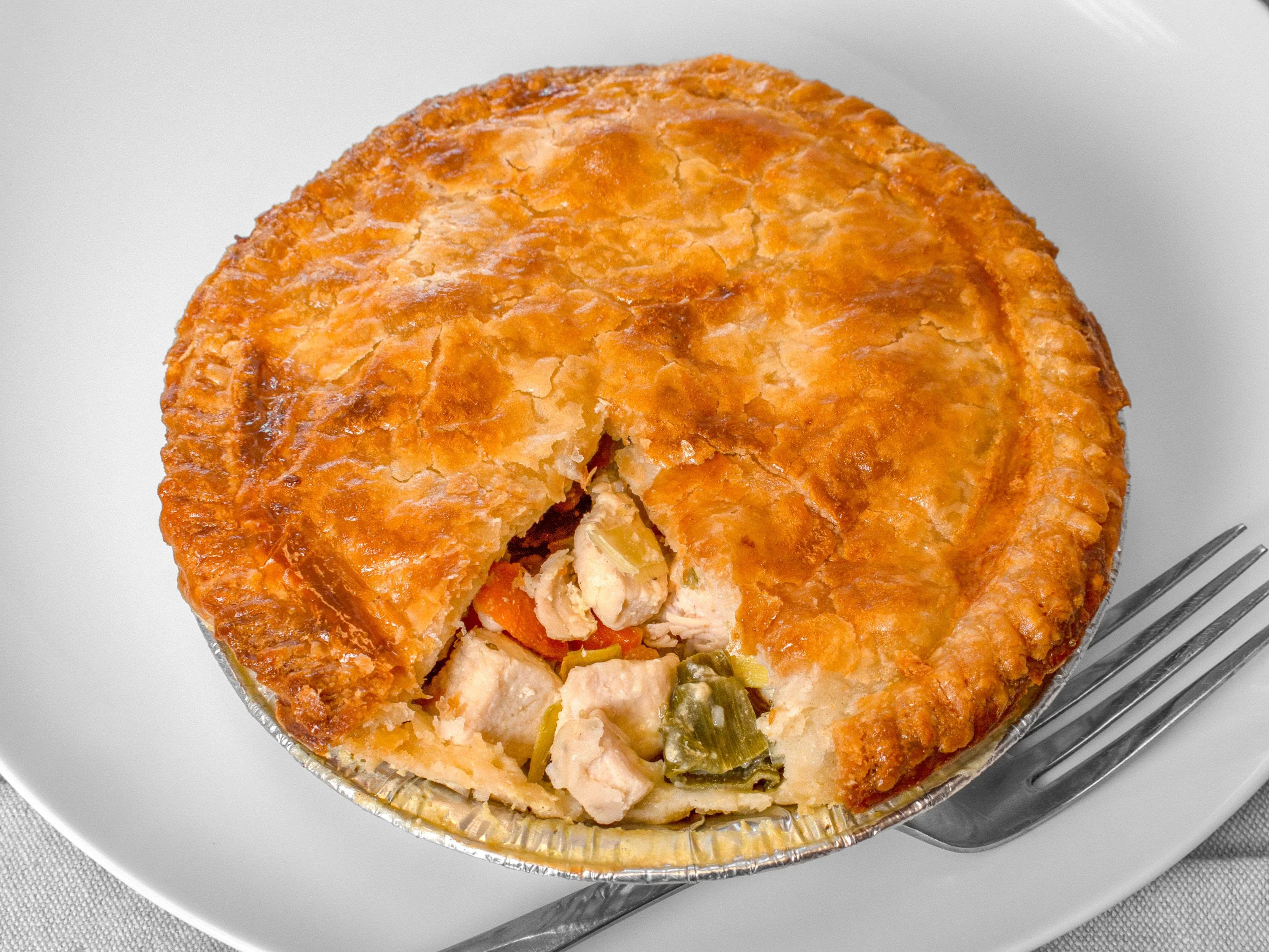 Golden flaky chicken pot pie in a foil tin with a wedge removed to reveal chunks of chicken and mixed vegetables, served on a white plate with a fork