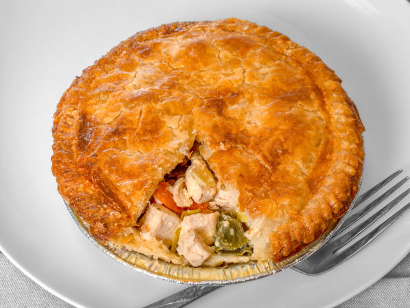 Golden flaky chicken pot pie in a foil tin with a wedge removed to reveal chunks of chicken and mixed vegetables, served on a white plate with a fork