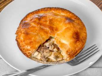 Comforting golden-brown flaky meat-and-vegetable pot pie in a foil tin with a wedge removed, revealing creamy filling, served on a white plate with a fork.