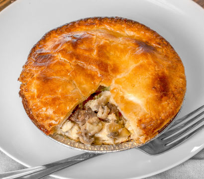 Comforting golden-brown flaky meat-and-vegetable pot pie in a foil tin with a wedge removed, revealing creamy filling, served on a white plate with a fork.
