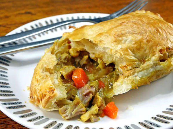 Golden flaky pastry hand pie filled with curried chicken, diced carrots and onions spilling onto a white patterned plate beside a fork