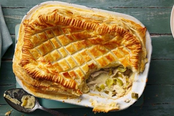 Golden puff pastry chicken and leek pie with creamy filling in a white baking dish on a teal wooden table, a serving spoon with sauce beside it
