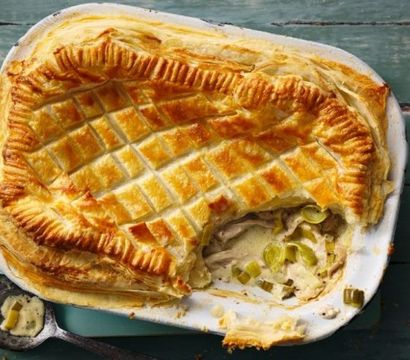 Golden puff pastry chicken and leek pie with creamy filling in a white baking dish on a teal wooden table, a serving spoon with sauce beside it