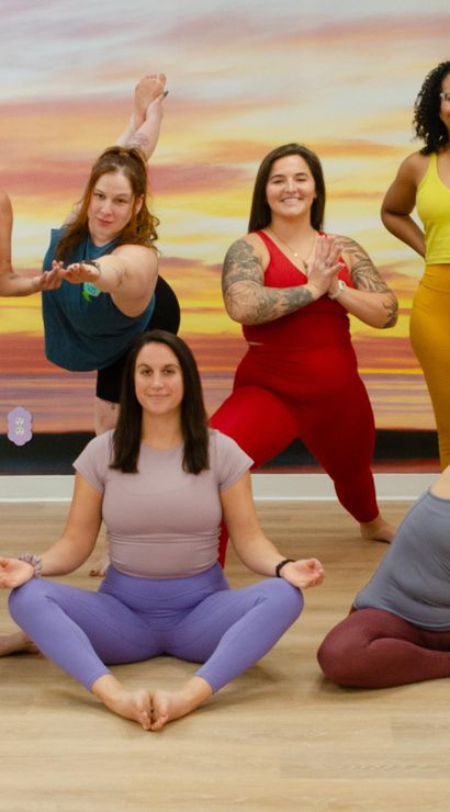 Seven women in colorful athletic wear practicing yoga poses in a bright indoor studio with a large sunset mural — seated meditation, side stretch, lunge and balancing poses in a friendly community yoga class.