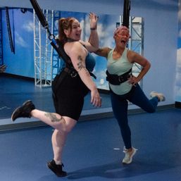 Two women wearing harnesses laughing and kicking during an indoor bungee aerial fitness class in front of a blue sky mural and mirrored wall