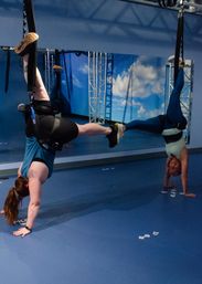 Aerial fitness class in a blue indoor studio: two people practicing inverted suspension training with harnessed legs in aerial straps while holding handstand-style poses in front of a sky mural.