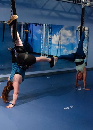 Aerial fitness class in a blue indoor studio: two people practicing inverted suspension training with harnessed legs in aerial straps while holding handstand-style poses in front of a sky mural.
