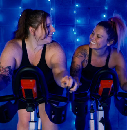 Two women smiling at each other while riding stationary bikes in a blue-lit indoor spin studio, wearing black workout tops and showing arm tattoos.