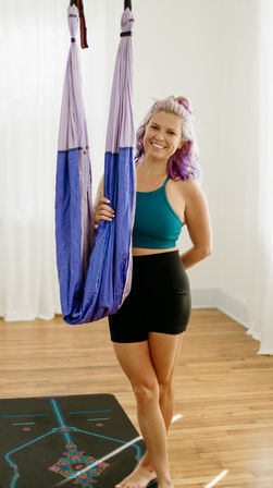 Smiling woman with purple hair in a teal sports bra and black shorts holding a purple aerial yoga hammock in a bright hardwood-floored yoga studio over a patterned yoga mat