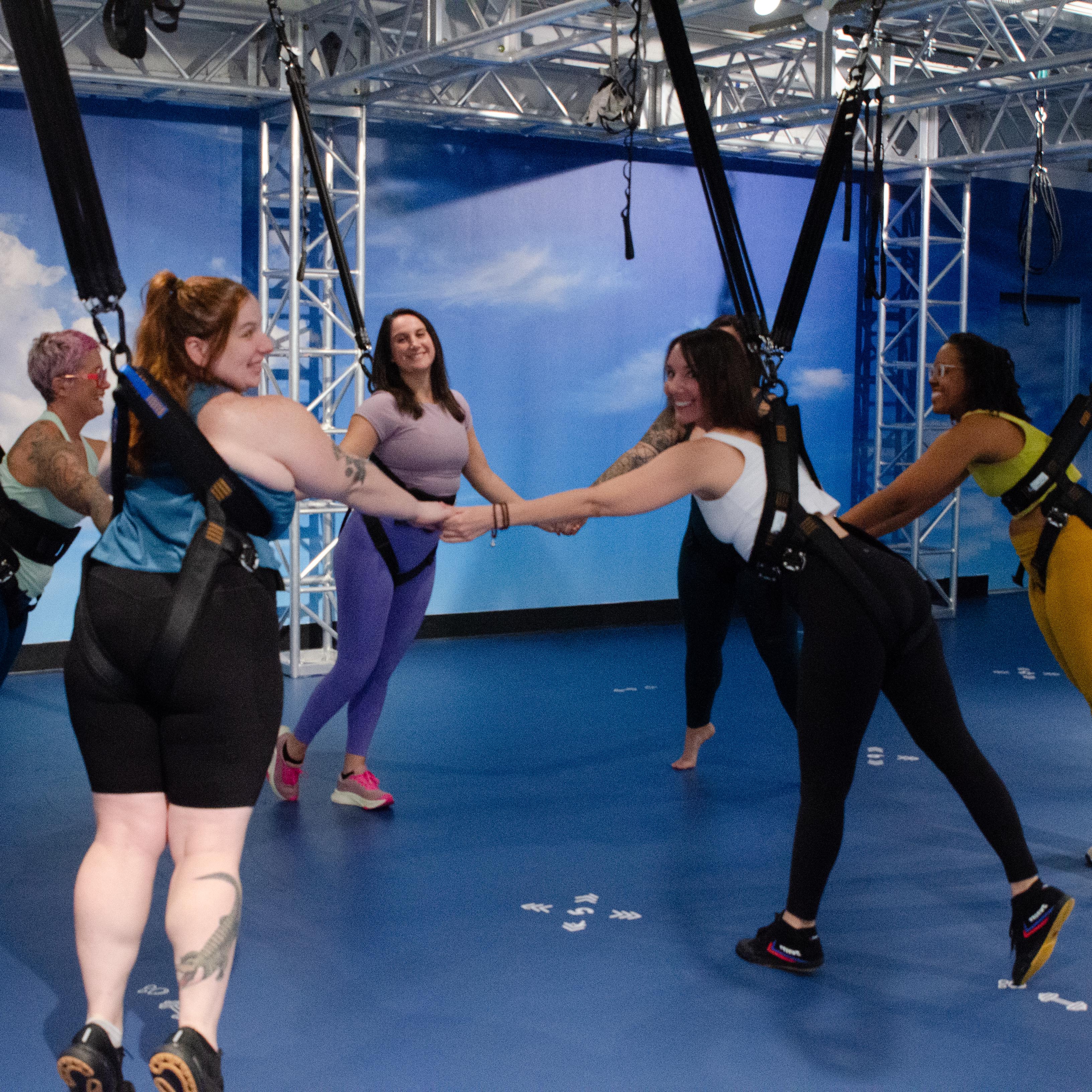 Group of women in a bungee fitness class wearing suspension harnesses, holding hands in a smiling circle during an indoor aerial fitness workout in a bright blue studio.