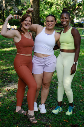 Three friends in colorful activewear smiling and posing in a sunny park, one flexing her bicep — outdoor group fitness photo with trees and grass