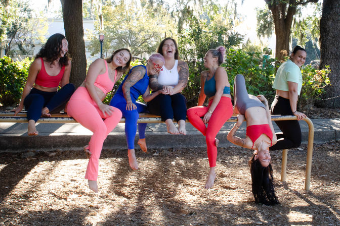 Group of seven women in colorful activewear laughing and playing on a park railing, one hanging upside down, with sunlit trees and wood-chip ground behind them.