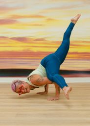 Grinning yogi with pink hair and red glasses in teal leggings performing an advanced arm-balance pose on a wood floor in a yoga studio in front of a vibrant sunset mural.