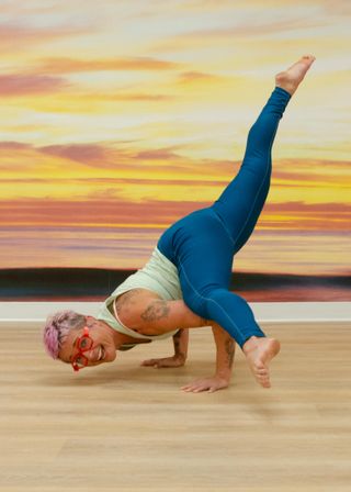 Grinning yogi with pink hair and red glasses in teal leggings performing an advanced arm-balance pose on a wood floor in a yoga studio in front of a vibrant sunset mural.