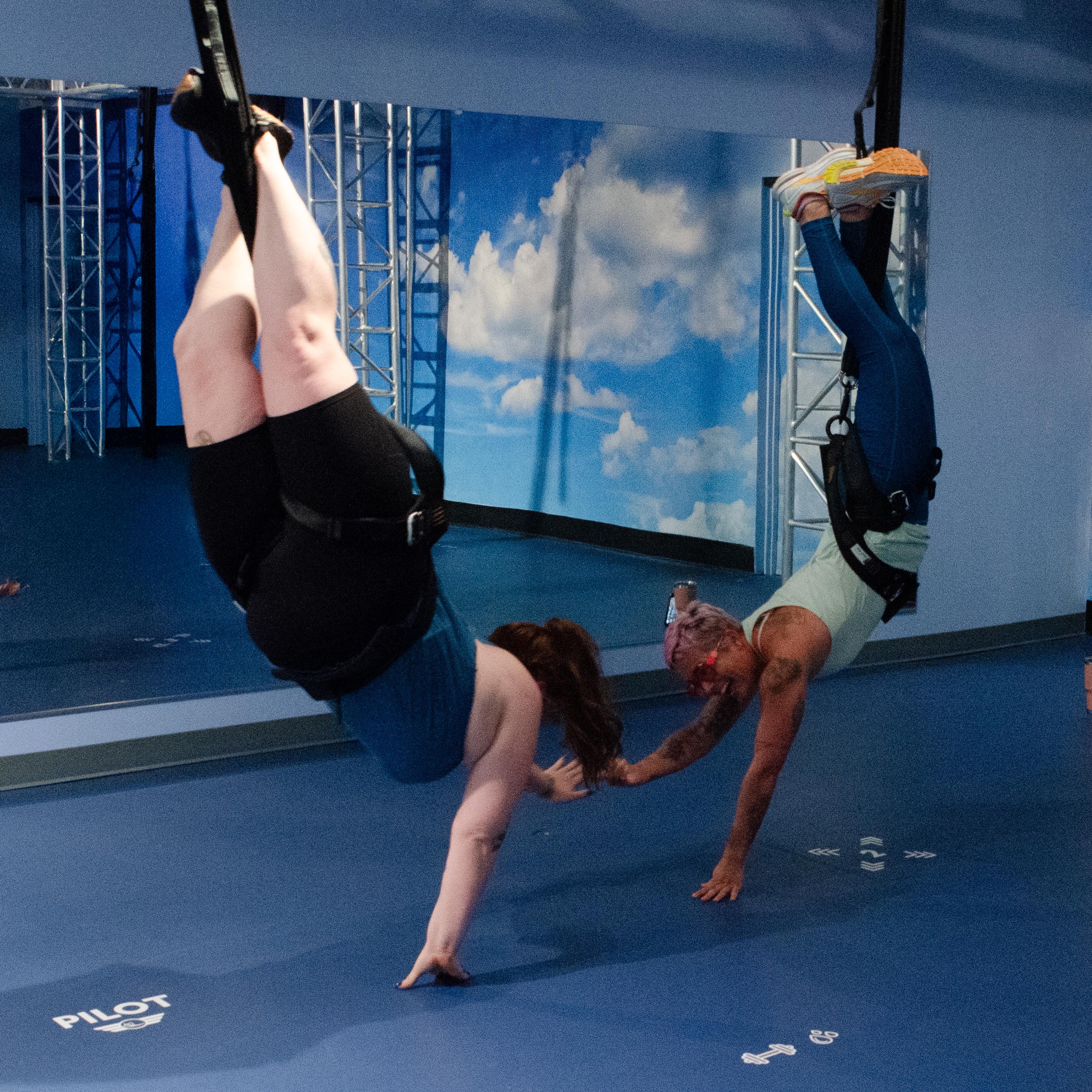 Two people inverted in harnesses reaching to touch hands during an aerial fitness class in an indoor studio with a blue padded floor, mirror wall, sky mural and metal rigging — suspension training/inversion workout.