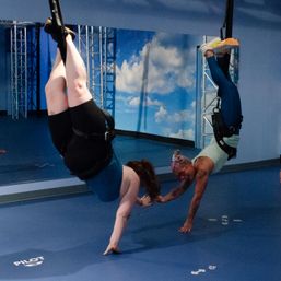 Two people inverted in harnesses reaching to touch hands during an aerial fitness class in an indoor studio with a blue padded floor, mirror wall, sky mural and metal rigging — suspension training/inversion workout.
