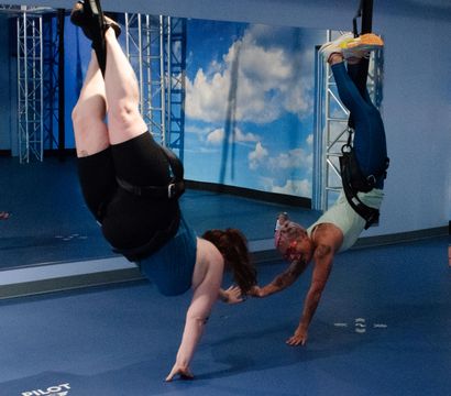 Two people inverted in harnesses reaching to touch hands during an aerial fitness class in an indoor studio with a blue padded floor, mirror wall, sky mural and metal rigging — suspension training/inversion workout.