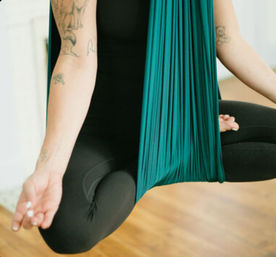 Close-up of a person practicing aerial yoga in a teal silk hammock, seated cross-legged in black leggings with tattooed arms and a meditative hand gesture over a wooden studio floor.