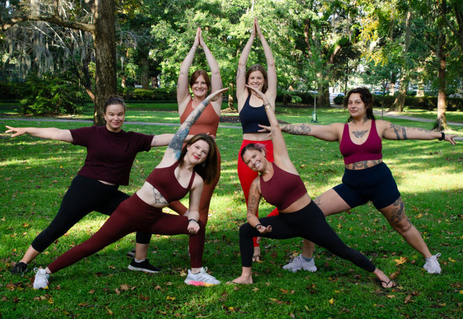 Six women in colorful activewear striking playful, synchronized yoga poses on a grassy, tree‑shaded public park lawn — outdoor group fitness and wellness session.