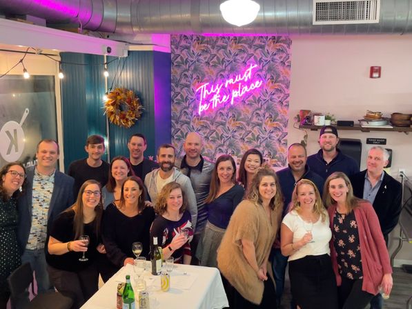 Large group of people posing inside a trendy indoor bar or restaurant with tropical wallpaper and a pink neon sign reading 'This must be the place', drinks on a table, string lights and exposed ductwork overhead.