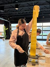 Smiling woman in an apron pulling a long sheet of fresh pasta from a hand-crank pasta machine during a hands-on cooking class in a bright industrial-style culinary studio.
