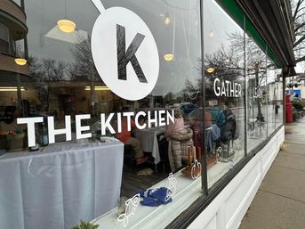 Urban cafe storefront window with bold white logo and lettering, rainy sidewalk reflections, diners seated at white-clothed tables and warm hanging lights visible inside.