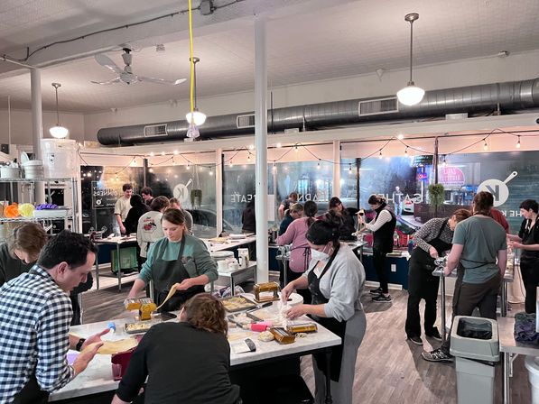 Hands-on pasta-making cooking class in a community culinary studio, people in aprons rolling dough and using pasta machines at shared worktables under string lights