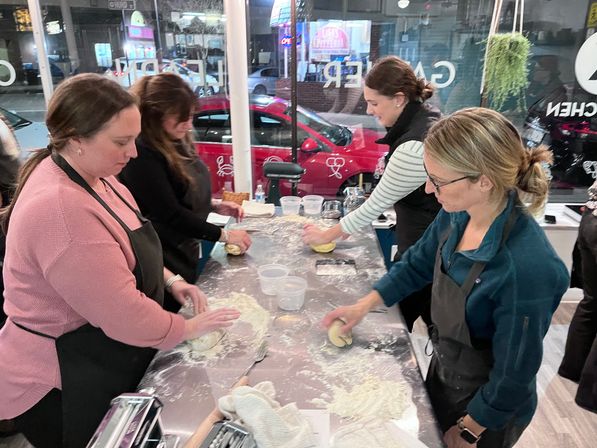 Four women in aprons shaping dough on a flour-dusted stainless table during a hands-on pasta-making class in a cozy urban kitchen studio with storefront windows and a red car visible outside.