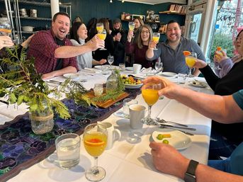Friends toasting mimosas at a bright local café brunch table with white linens, a greenery centerpiece, fruit plates and coffee