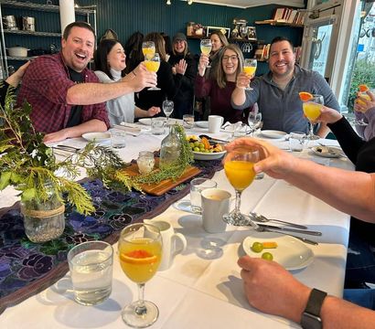 Friends toasting mimosas at a bright local café brunch table with white linens, a greenery centerpiece, fruit plates and coffee