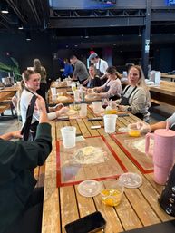 Group baking class at a long wooden table in an indoor culinary studio, participants rolling dough on silicone mats with eggs, measuring cups and drinks nearby