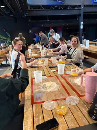Group baking class at a long wooden table in an indoor culinary studio, participants rolling dough on silicone mats with eggs, measuring cups and drinks nearby