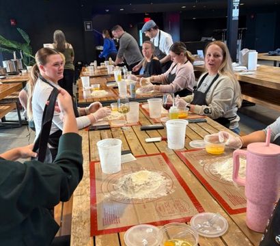Group baking class at a long wooden table in an indoor culinary studio, participants rolling dough on silicone mats with eggs, measuring cups and drinks nearby