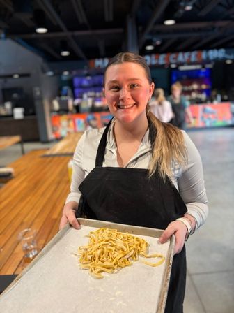 Smiling person in a black apron holding a baking sheet of freshly made fettuccine pasta in a modern cafe with wooden communal tables