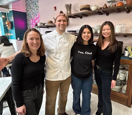 Smiling chef in white jacket poses with three guests in a cozy urban cafe interior with open shelves of dishes, a pink neon sign and a marble table.