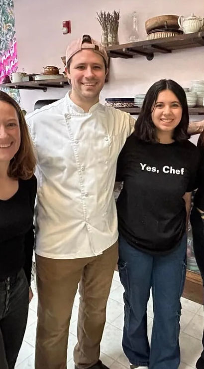 Smiling chef in white jacket poses with three guests in a cozy urban cafe interior with open shelves of dishes, a pink neon sign and a marble table.