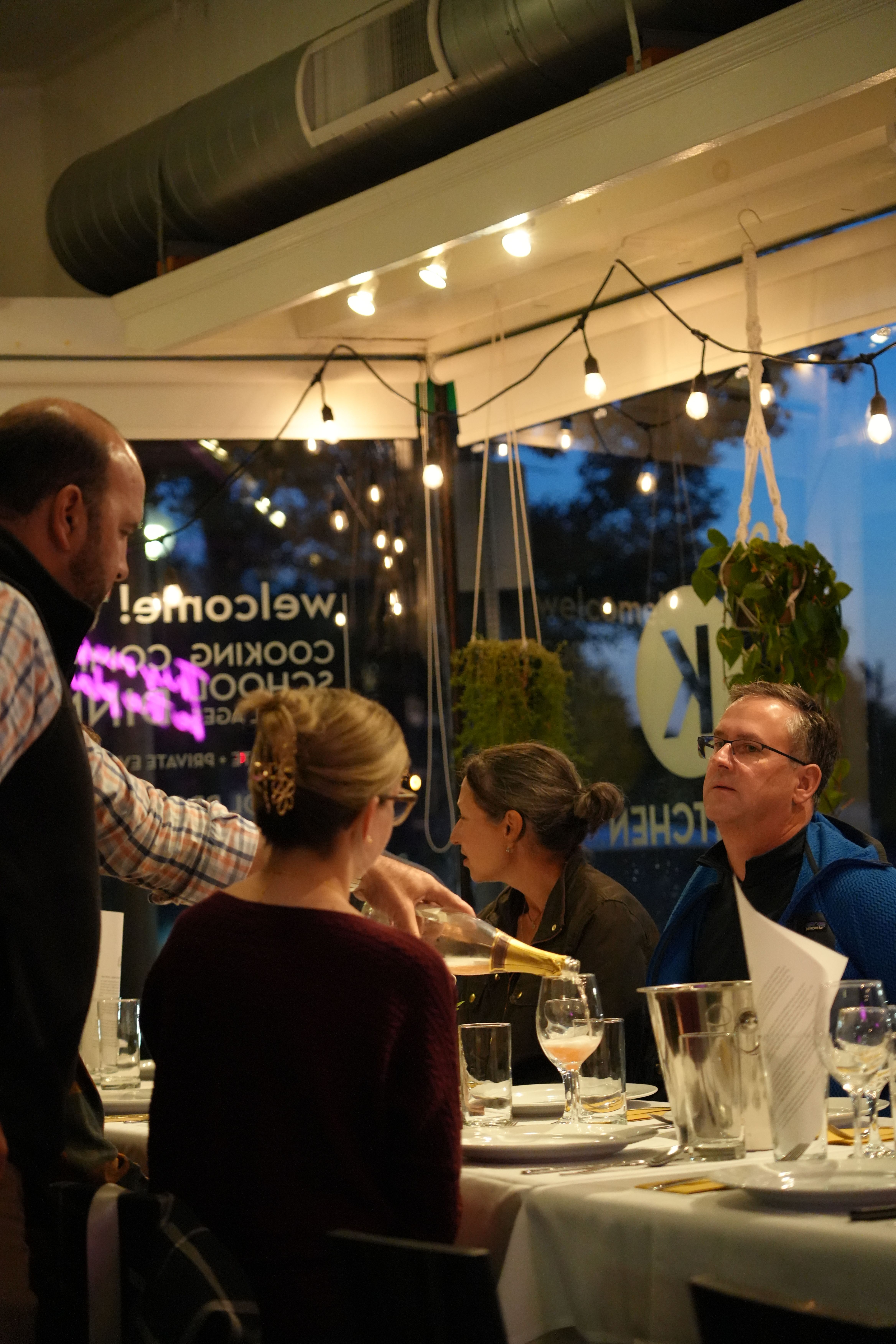 Evening dinner at a cozy restaurant: a server pours sparkling wine for seated adults under warm string lights and hanging plants.