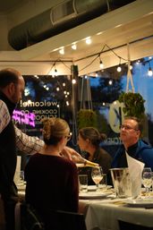 Evening dinner at a cozy restaurant: a server pours sparkling wine for seated adults under warm string lights and hanging plants.