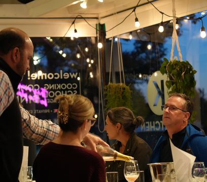Evening dinner at a cozy restaurant: a server pours sparkling wine for seated adults under warm string lights and hanging plants.