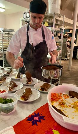 Chef in a commercial kitchen giving the finishing touch, spooning warm brown sauce over rolled savory enchiladas on small white plates at a busy prep station with garnishes and serving bowls.